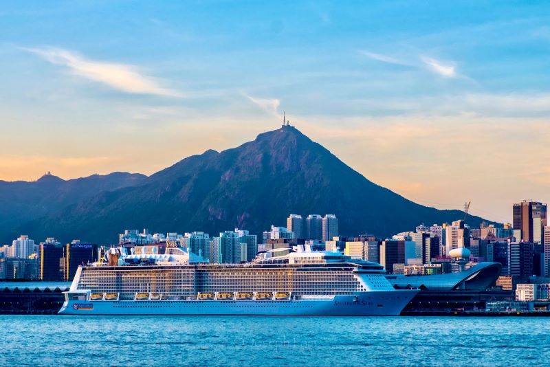 Het Royal Caribbean-cruiseschip Spectrum of the Seas ligt bij schemering afgemeerd in een haven, met op de achtergrond een levendige skyline en een berg, onder een lucht die overgaat in de avond.